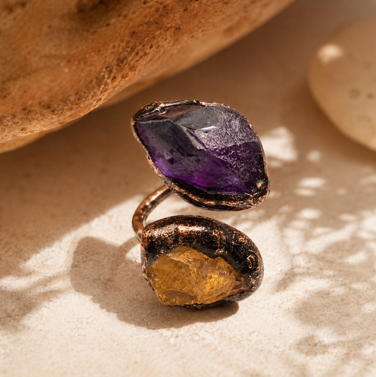 Purple yellow gemstone ring displayed on warm stone with bold two-stone contrast and artistic styling.