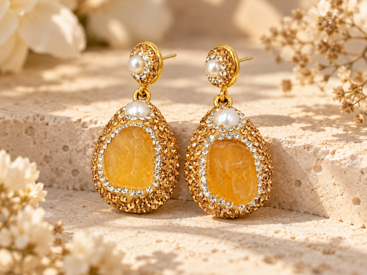 Pair of honey-yellow citrine drop earrings with white pearl accents and ornate gold-tone borders on a sunlit cream stone surface