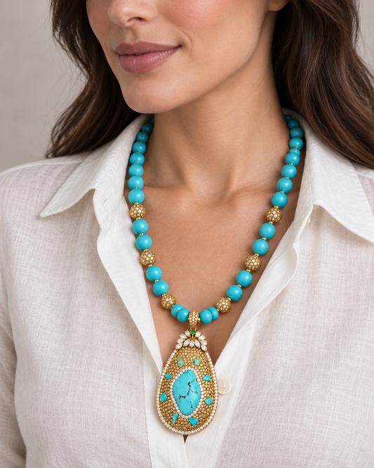 Angled close-up of a turquoise pendant and beaded necklace worn with an ivory shirt, showing gold-tone details.