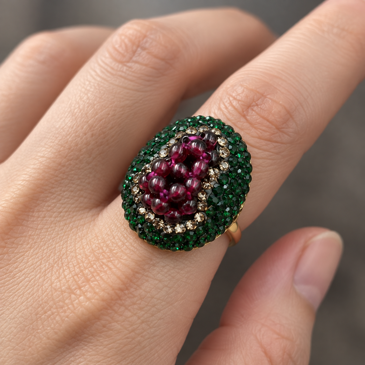 Close-up of a garnet cocktail ring on an index finger, showing wine-red beads, a green crystal border and a gold-tone band.