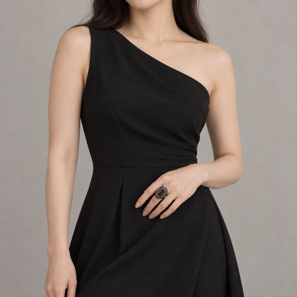 Woman wearing a black one-shoulder dress with a wine-red and green statement ring on her hand near her waist.