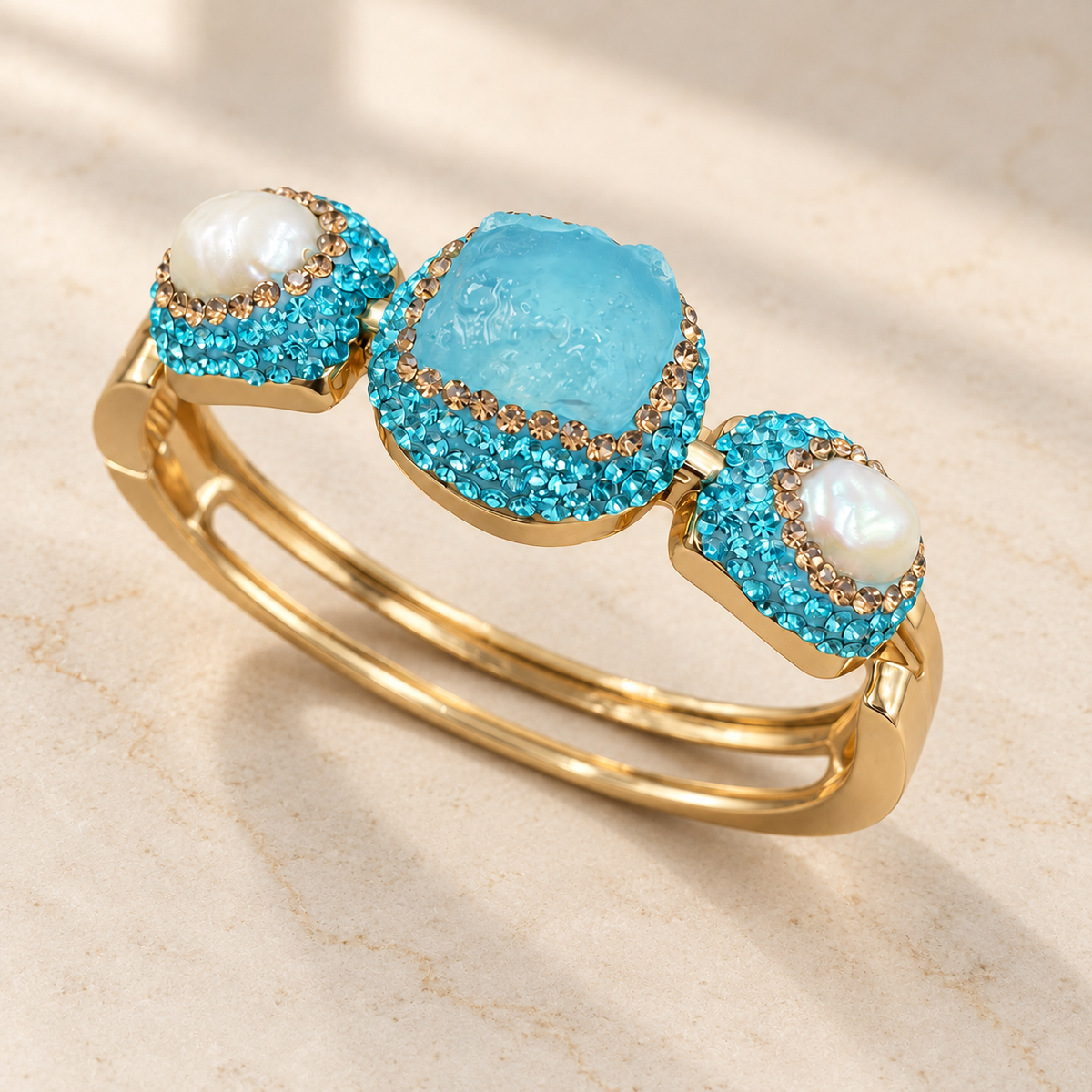 Angled view of a turquoise bangle with freshwater pearls, sparkling crystal borders and twin gold-tone bands on an ivory stone surface.