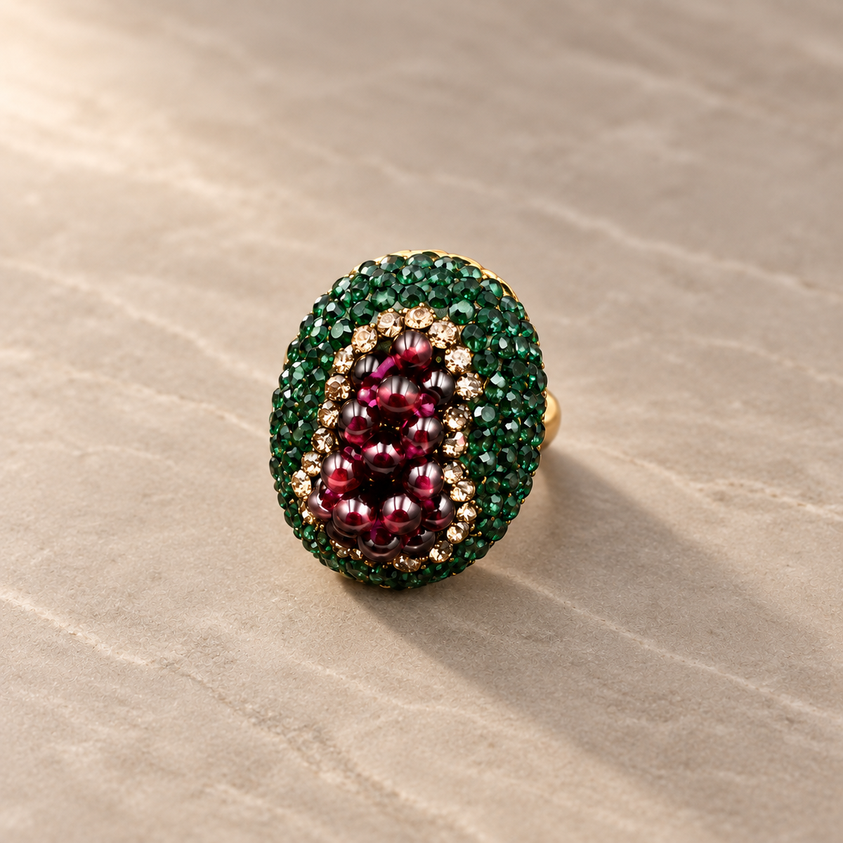 Wine-red garnet cocktail ring with green crystal accents standing at a slight angle on a smooth beige stone surface in soft side light.