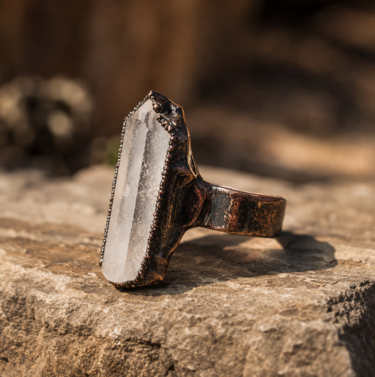 Clear quartz point ring with a transparent crystal column, sculptural setting, and natural stone background.