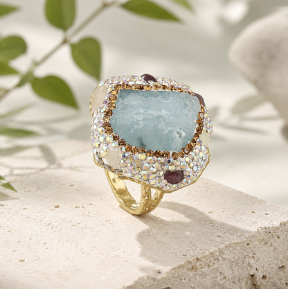 Aquamarine ring with raw seafoam-blue crystal center, gold-tone band, and sparkling accents on a natural stone background.