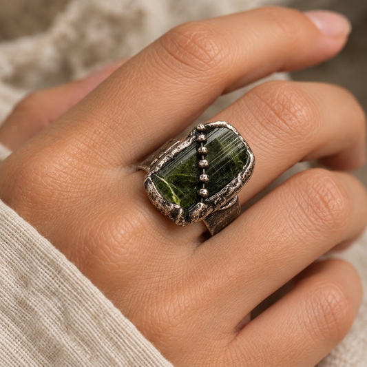 Green tourmaline ring worn on the hand, showing the elongated raw stone and distressed metal setting.