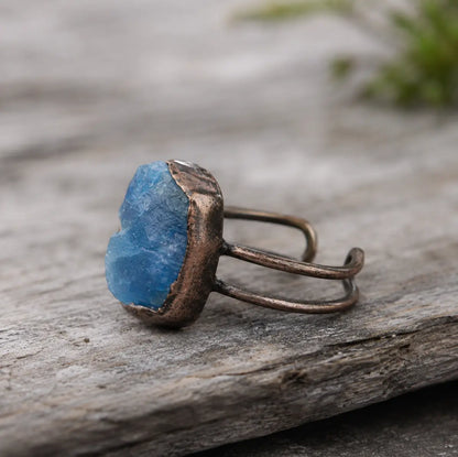 Blue apatite ring with raw lake-blue gemstone, organic open band, and natural stone background.
