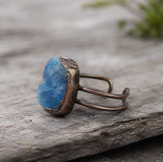 Blue apatite ring with raw lake-blue gemstone, organic open band, and natural stone background.