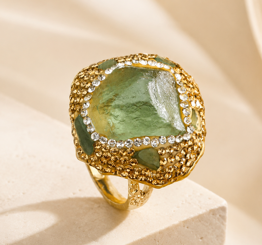 Green fluorite ring with raw green crystal center, gold-tone setting, and sparkling accents on a soft neutral background.
