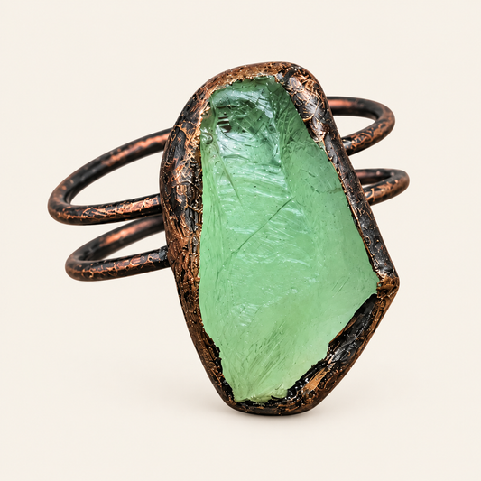 A handmade ring featuring a large, raw green stone in a textured, dark bronze bezel setting with a wrap-around coppery metal band, shown on a light cream background.