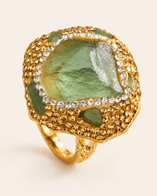 Green fluorite ring with an irregular translucent stone, clear decorative accents and a gold-tone band on a warm off-white background