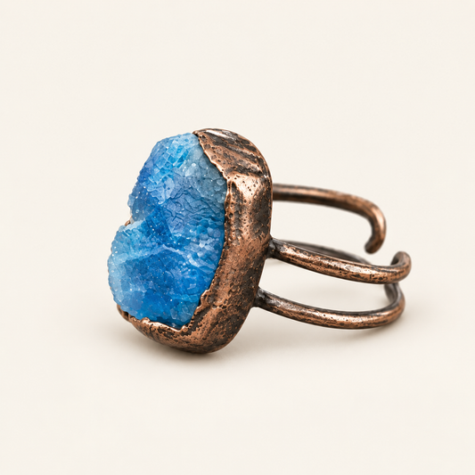 A rustic open-band ring featuring a rough, textured blue stone set in a hammered copper-toned metal, photographed on a clean, solid off-white background.