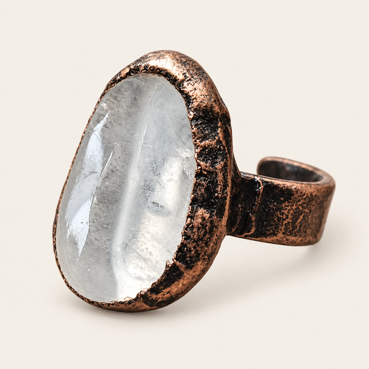 Polished clear quartz crystal ring on warm off-white, showing the translucent stone and broad dark-toned setting.