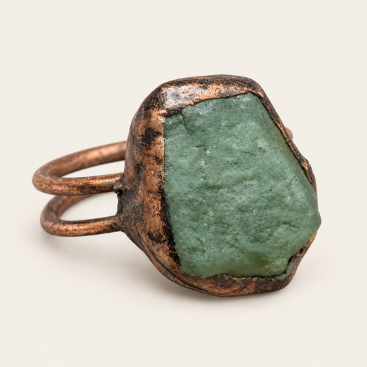 A handmade ring featuring a large, rough green stone in a textured, rustic copper-toned bezel setting with a double-band design, shown on a light beige background.