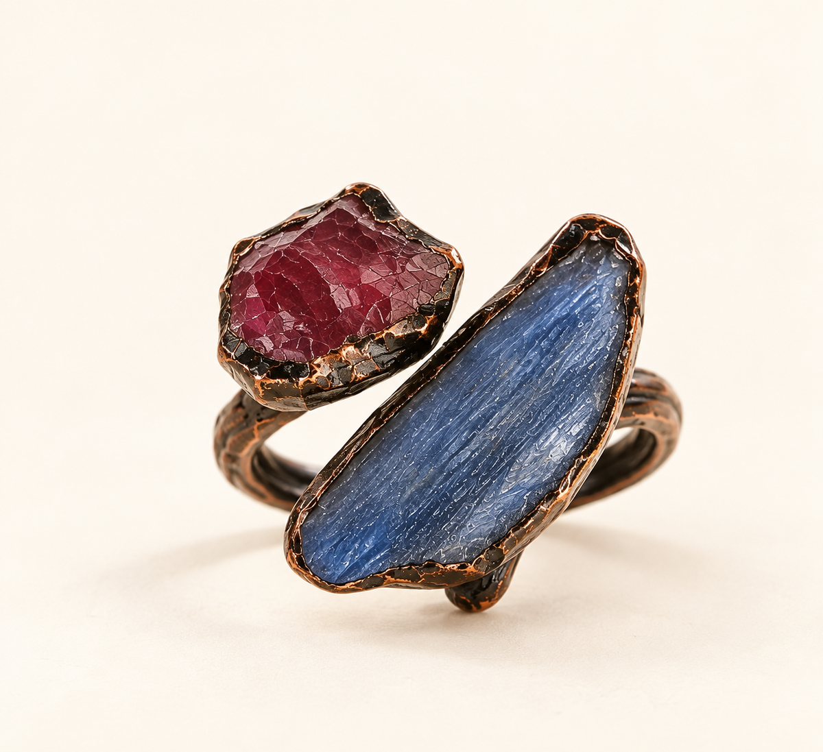 Crimson Tide red stone and blue kyanite ring with an asymmetrical two-stone design on an off-white background.