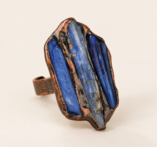 Front view of a blue kyanite three-stone ring with elongated raw stones and an open copper band on an off-white background.