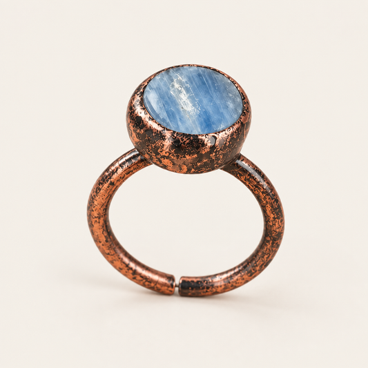 Front view of a blue apatite ring with a round raw stone, visible surface textures and an open band on an off-white background.