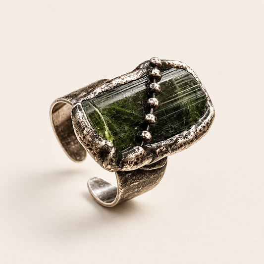 Deep olive green tourmaline ring on warm off-white, showing raw striations, raised setting, and open band.