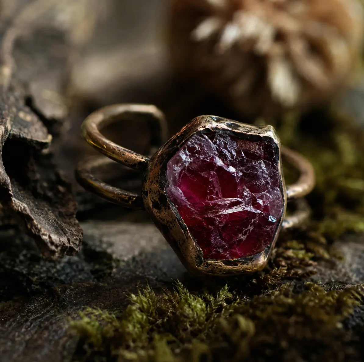 Red gemstone ring displayed on dark natural wood with moss, earthy texture, and refined depth.
