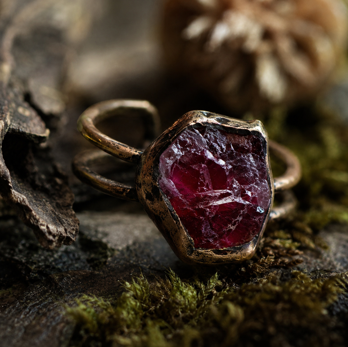 Red gemstone ring displayed on dark natural wood with moss, earthy texture, and refined depth.