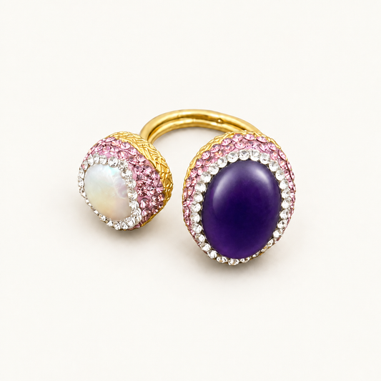 Amethyst and baroque pearl ring with pink and clear pavé crystal halos and a gold-tone open band on an off-white background.