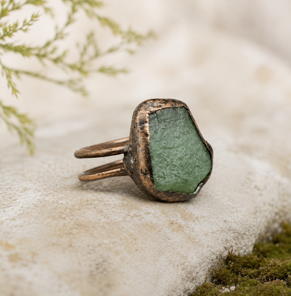 Apatite ring displayed on pale stone with soft greenery, earthy texture, and refined styling.